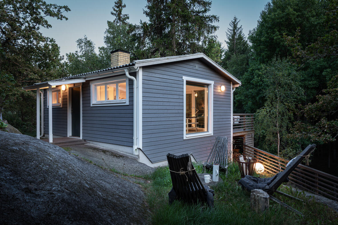 Small grey timber cabin illuminated at twilight and surrounded by forest. Small grey timber cabin illuminated at twilight and surrounded by forest.
